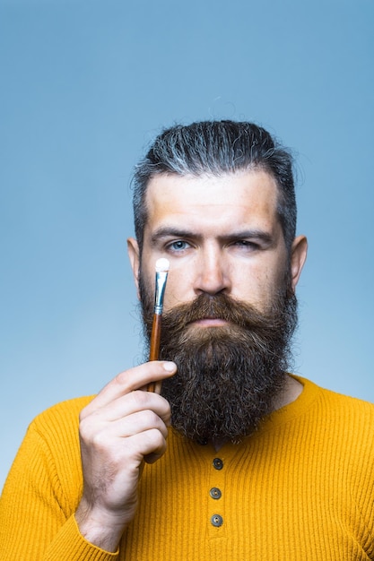 Bonito homem barbudo com barba longa exuberante e bigode no rosto sério, segurando o pincel na camisa amarela no estúdio sobre fundo azul