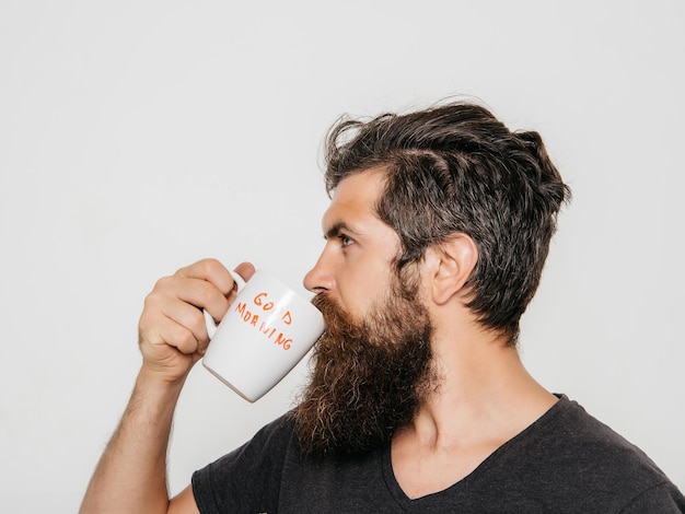 Bonito homem barbudo com cabelo estiloso, barba e bigode no rosto sério na camisa segurando o copo branco