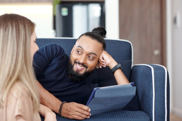 Bonito homem barbudo com o cabelo preso em um coque, descansando no sofá com o computador tablet e olhando para a esposa contando uma piada engraçada