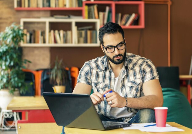 Bonito homem barbudo com óculos trabalhando com laptop na moderna e elegante sala de coworking com conceito de sucesso de pensamento inteligente de estantes