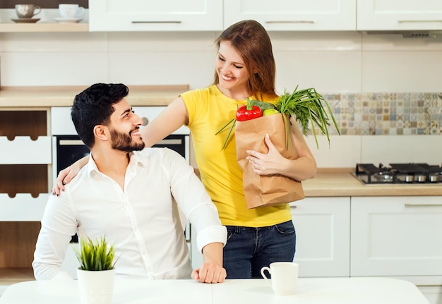 Bonito homem barbudo de camisa branca imaculada sentado à mesa mulher alta e magra em camiseta amarela ao lado do marido segurando sacola de compras com alimentos nutritivos saudáveis