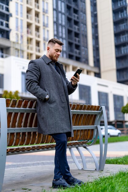 Bonito homem barbudo de casaco cinza olha para a tela do telefone enquanto se inclina no banco Edifício moderno ao fundo