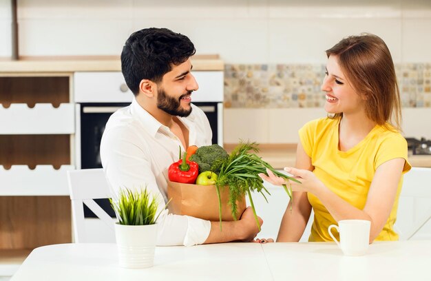 Bonito homem barbudo e mulher magra sentada à mesa segurando sacola de compras com alimentos nutritivos saudáveis