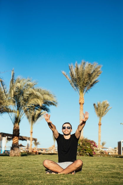 Bonito homem barbudo em óculos de sol, sentado na grama verde e relaxe, aproveite as férias de verão com as mãos levantadas, sorriso feliz.