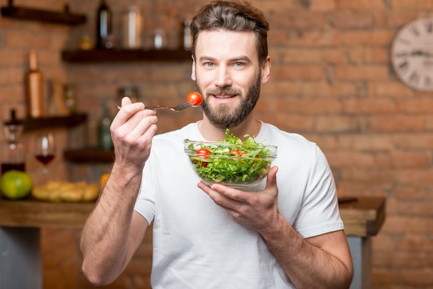 Bonito homem barbudo em t-shirt branca comendo salada com tomates na cozinha. Conceito de comida saudável e vegana