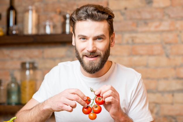 Bonito homem barbudo em t-shirt branca fazendo salada com tomate na cozinha. Conceito de comida saudável e vegana