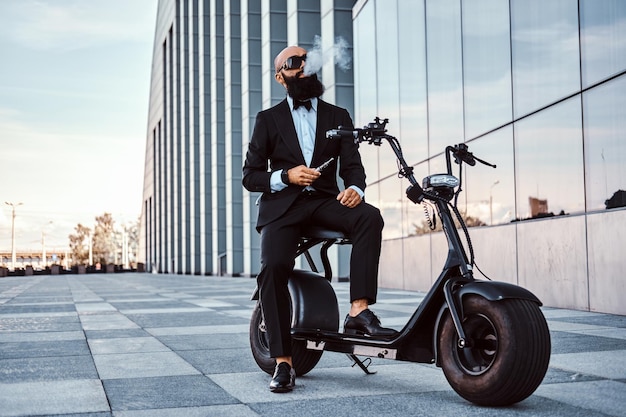 Bonito homem barbudo está fazendo uma pequena pausa enquanto fuma vaporizador. homem está sentado na scooter elétrica.