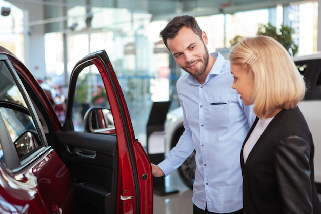 Bonito homem barbudo falando com sua esposa ao escolher o carro novo para comprar na concessionária