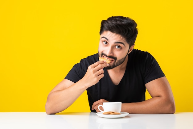 Bonito homem barbudo indiano comendo biscoitos integrais saudáveis mergulhados em um café de chá ou chai em uma xícara, sentado à mesa contra um fundo amarelo