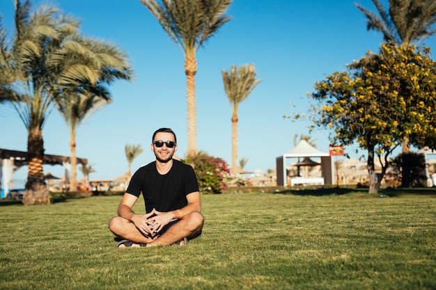 Bonito homem barbudo magro em óculos de sol, sentado na grama verde e relaxe aproveite as férias de verão.