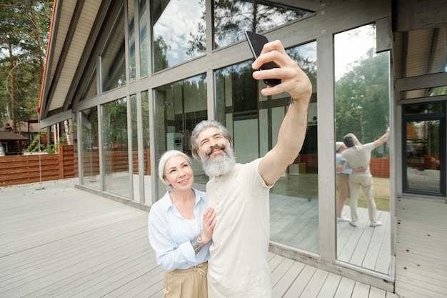 Bonito homem barbudo sênior abraçando a esposa enquanto tirava uma selfie com ele perto da casa de vidro
