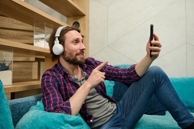 Bonito homem caucasiano barbudo relaxado usando fones de ouvido sem fio falando por videochamada olhando para a câmera web no smartphone na mão relaxando deitado em um sofá em casa