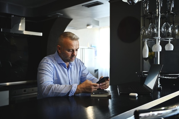 Bonito homem caucasiano de meia idade, freelancer, empresário de sucesso na camisa azul casual, usando o telefone celular enquanto trabalhava online, sentado no bar em sua cozinha moderna em casa.