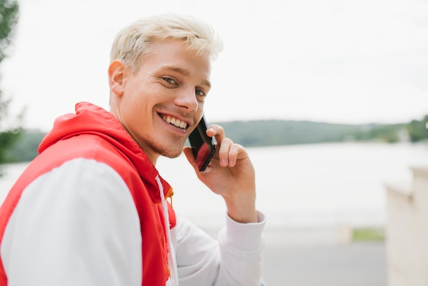 Bonito homem caucasiano loiro sorrindo enquanto conversava com seu amigo no telefone inteligente em pé no parque Comunicação de pessoas viajando e conceito de tecnologia moderna