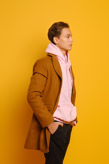 Bonito homem elegante em trenchcoat marrom e moletom rosa com capuz em perfil sobre fundo amarelo do estúdio