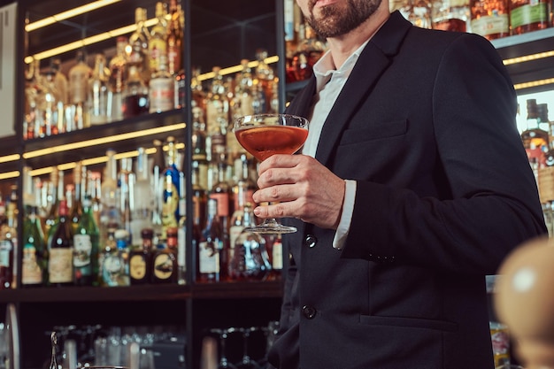 Bonito homem elegante segura um copo de álcool exclusivo em pé no fundo do balcão de bar.