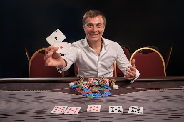 Bonito homem emocional em uma camisa branca está jogando poker sentado à mesa no cassino. Ele está fazendo apostas esperando uma grande vitória e mostrando suas cartas. Apostas por dinheiro. Jogos de fortuna.