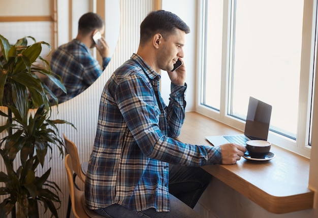 Bonito homem freelancer falando no telefone e trabalhando remotamente no laptop e tomando café no café