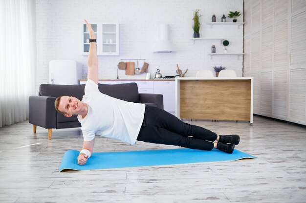 Bonito homem musculoso com uma camiseta está fazendo exercícios funcionais no chão em casa. Fitness em casa. Estilo de vida saudável.