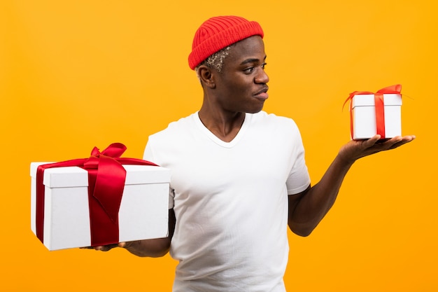 Bonito homem negro africano surpreso com um sorriso em uma camiseta branca detém duas caixas de presente com uma fita vermelha para um aniversário em um estúdio amarelo