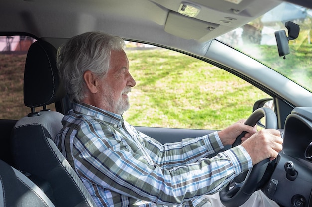 Bonito homem sênior com camisa quadriculada, sentado dentro de seu carro sorri enquanto olha para a frente Sorrindo avô idoso barbudo dirigindo o automóvel com as mãos no volante