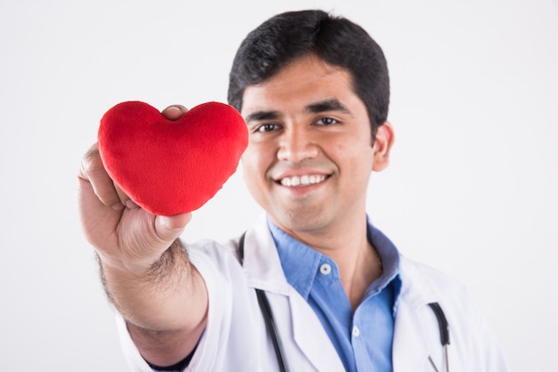 Bonito indiano masculino médico segurando o brinquedo vermelho de pelúcia como um conceito de cuidado cardíaco. de pé isolado sobre fundo azul