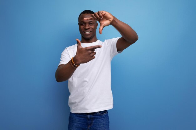 Bonito jovem africano em camiseta branca mostrando gesto fotográfico em fundo azul com espaço de cópia