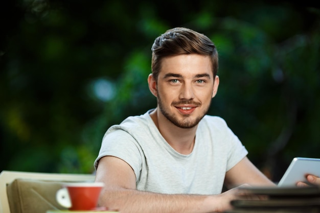 Bonito jovem alegre sentado à mesa no café ao ar livre segurando o tablet olhando na câmera