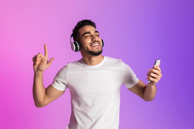 Bonito jovem árabe ouvindo música em fones de ouvido usando o novo aplicativo de smartphone e dançando em