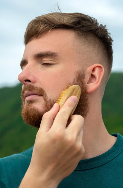 Bonito jovem barbudo homem brutal com cabelo de barba está penteando a barba no rosto com uma escova de pente Acessório de produtos de autocuidado pessoal masculino Foto vertical de fundo de verão natural