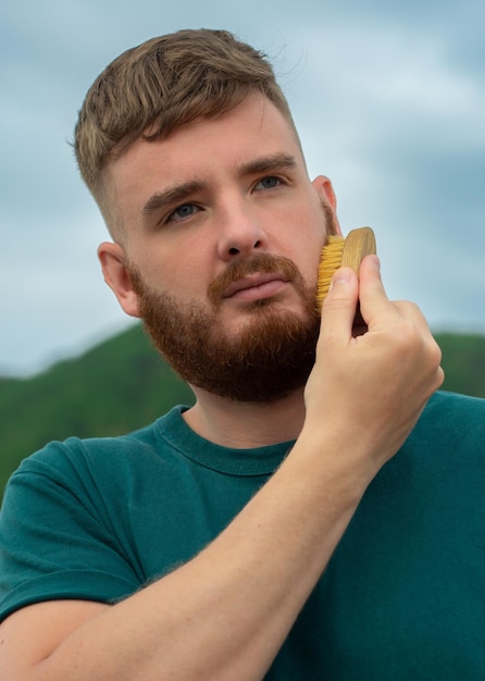 Bonito jovem barbudo homem brutal com cabelo de barba está penteando a barba no rosto com uma escova de pente Acessório de produtos de autocuidado pessoal masculino Foto vertical de fundo de verão natural