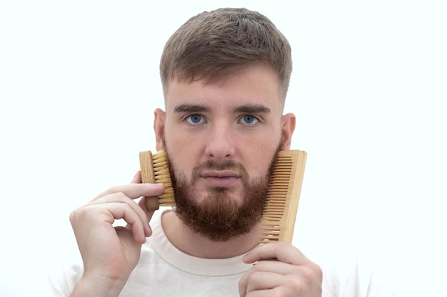 Bonito jovem barbudo Homem brutal europeu com cabelo de barba está penteando a barba no rosto com uma escova de pente Acessório de produtos de autocuidado pessoal masculino isolado no fundo branco