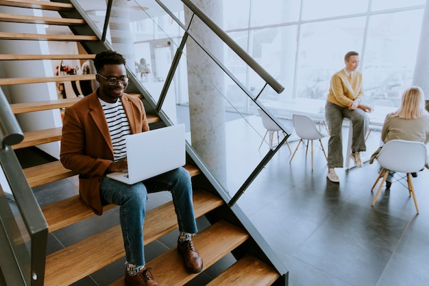 Bonito jovem empresário afro-americano trabalhando no computador portátil enquanto está sentado em uma escada de escritório