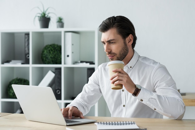 Bonito jovem empresário com café de papel trabalhando com laptop