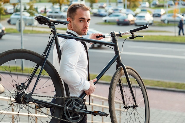 Bonito jovem empresário em uma camisa branca e gravata preta leva sua bicicleta