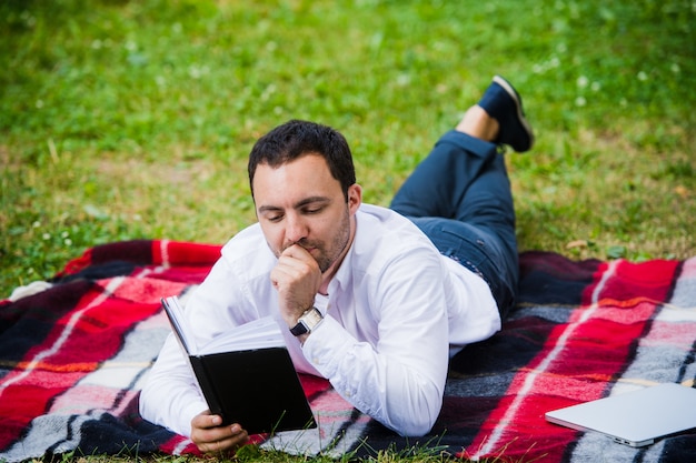 Bonito jovem empresário lendo livro deitado no parque
