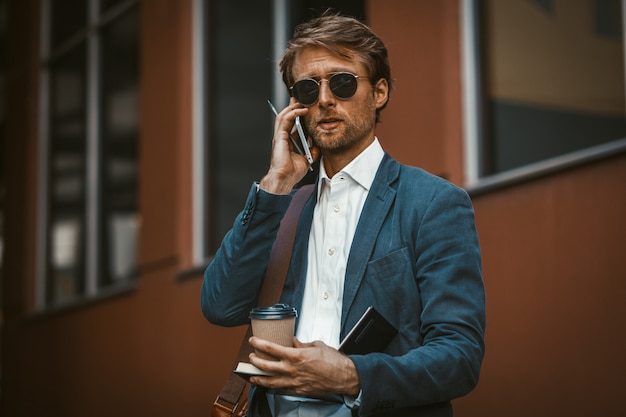 Bonito jovem empresário segurando copo descartável e falando ao telefone