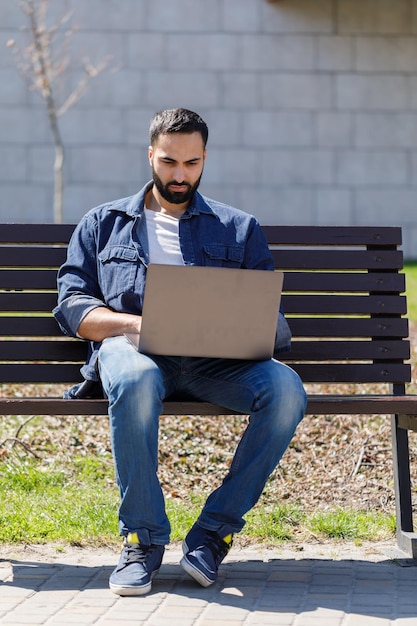 Bonito jovem empresário sentado em um banco com seu laptop em uma rua ao lado de um parque