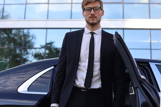 Foto bonito jovem empresário sério em pé perto de seu confortável carro novo e indo para o escritório.