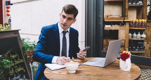 Bonito jovem empresário vestindo terno azul elegante e gravata listrada olhando de lado enquanto está sentado no terraço do café e trabalhando com laptop