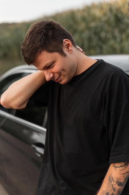 Bonito jovem hipster em camiseta preta e calça fica e posa perto de um carro cinza ao ar livre