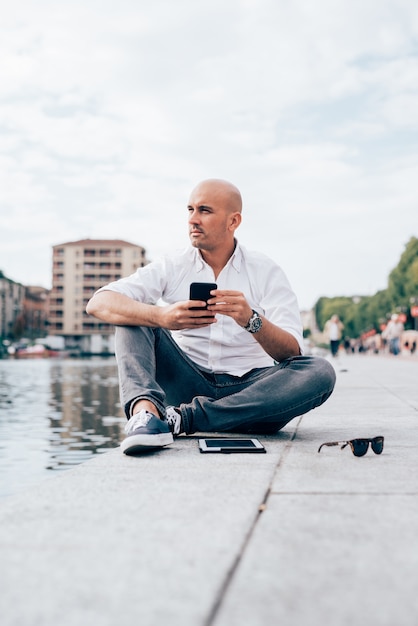 Bonito, jovem, homem negócios, em, um, camisa branca, sentada, por, a, água, usando, smartphone