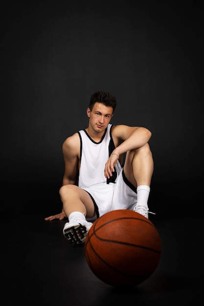 Bonito jovem jogador de basquete em roupas esportivas brancas senta-se no chão posando com uma bola isolada em fundo preto