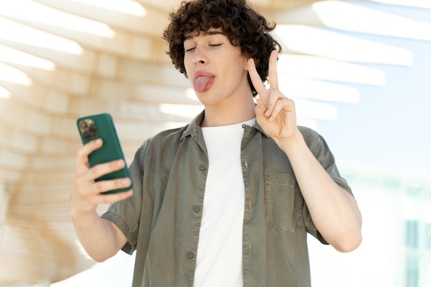 Bonito jovem macho com cabelo encaracolado em roupas elegantes tomando selfie no smartphone