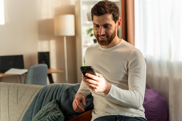 Bonito jovem macho sorrindo enquanto está sentado com o smartphone e olhando para a tela com cara de feliz. Foto