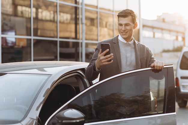 Bonito jovem moderno com telefone entrando em seu carro em pé ao ar livre