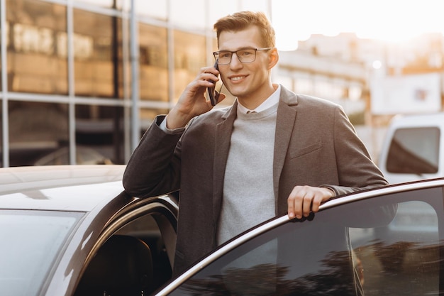 Bonito jovem moderno em óculos falando por telefone e entrando em seu carro em pé ao ar livre