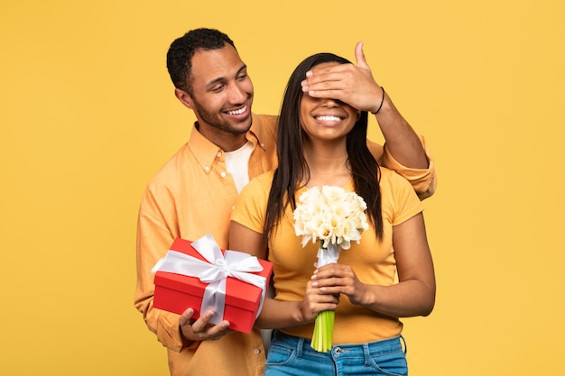 Bonito jovem negro dando flores e caixa de presente para sua namorada cobrindo os olhos dela fazendo