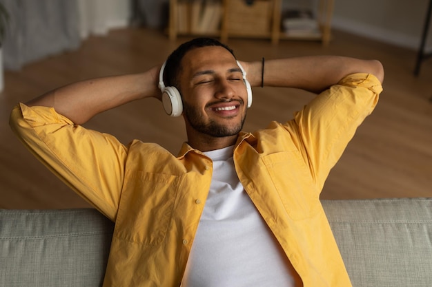 Bonito jovem negro em fones de ouvido relaxando no sofá com os olhos fechados curtindo música em casa