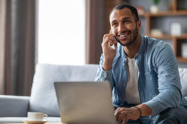 Bonito jovem negro falando no celular e usando o laptop em casa